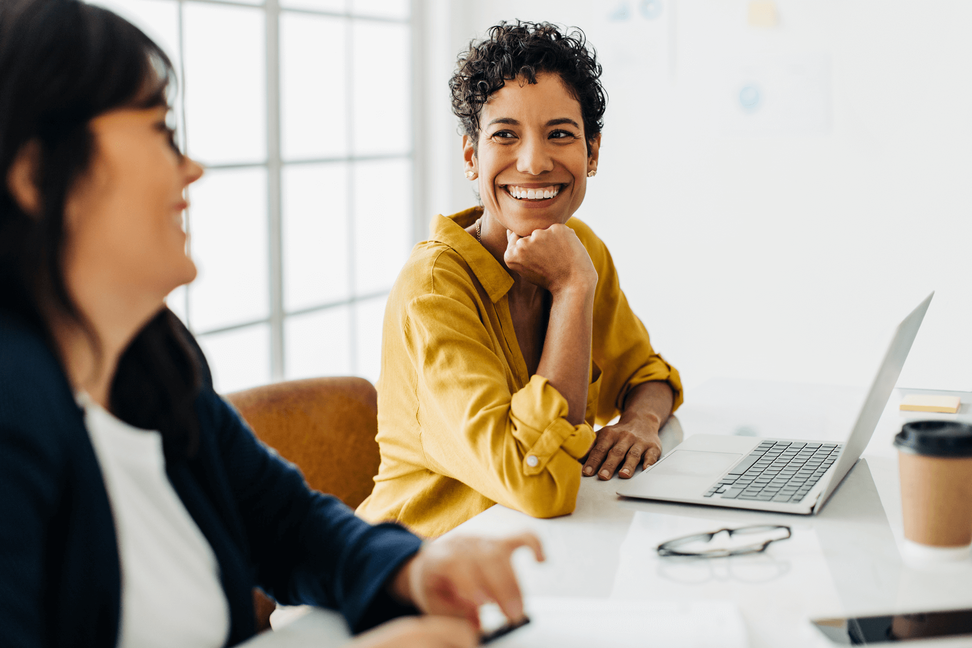 Two women sitting at a desk laughing with a computer in front of one woman